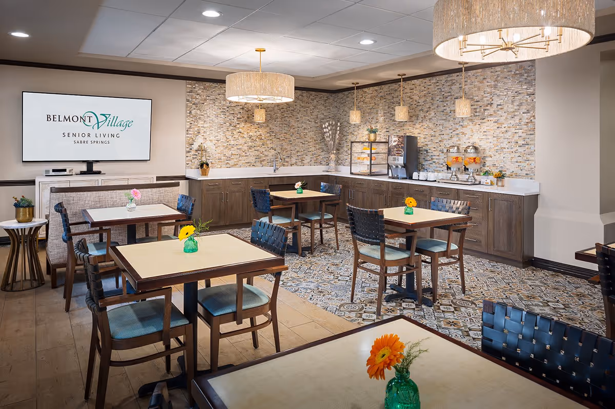 A bright and modern dining area in Belmont Village Senior Living Sabre Springs featuring several square tables with chairs, each table decorated with a small green vase holding a single colorful flower. The back wall has a mosaic tile backsplash with wooden cabinets below and a countertop holding beverage dispensers and a coffee machine. Two large pendant lights hang from the ceiling, and a flat screen on the left wall displays the Belmont Village Senior Living Sabre Springs logo.