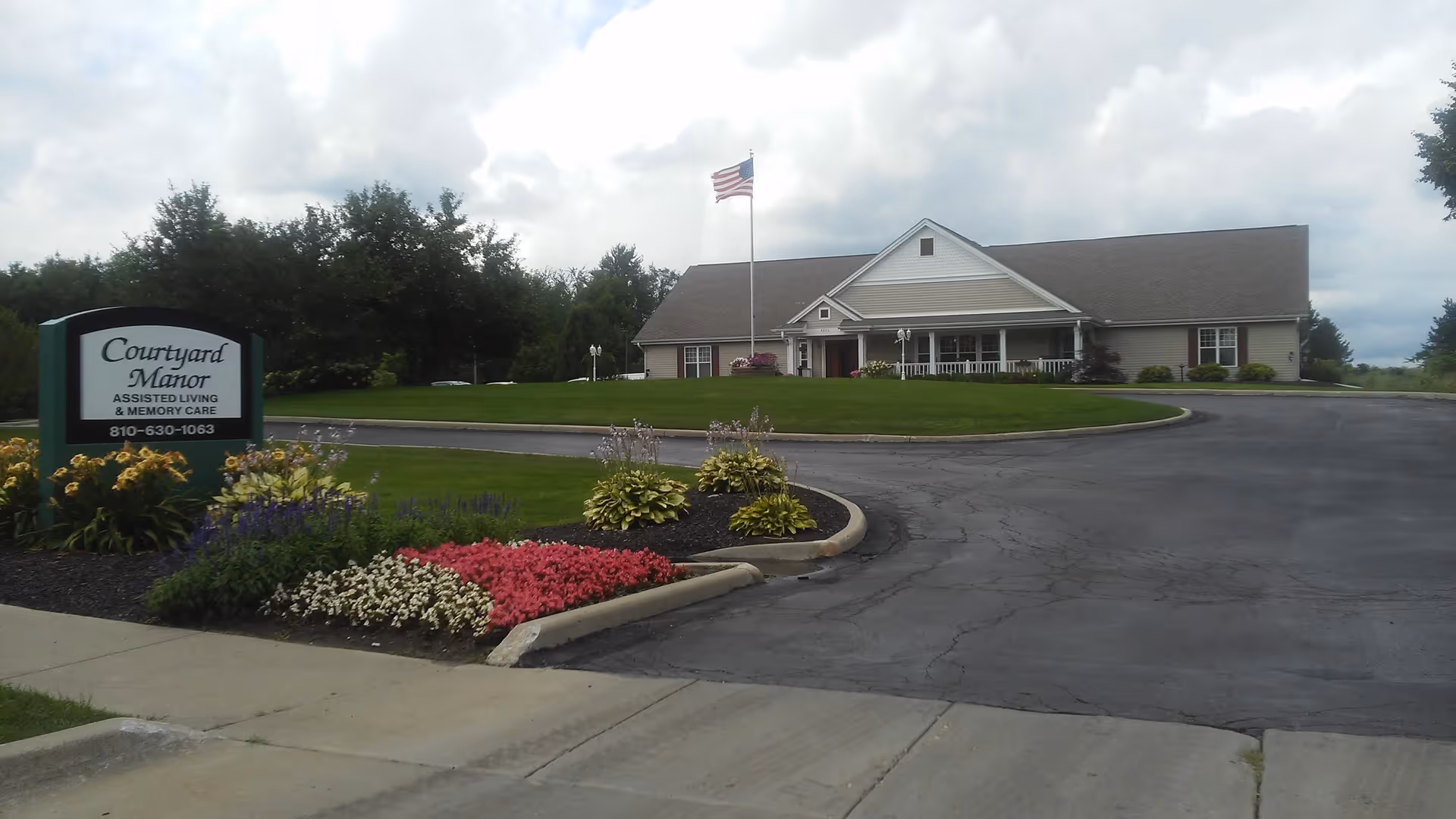Exterior view of Courtyard Manor of Swartz Creek, an assisted living and memory care facility. The image shows a single-story building with a gabled roof, a front porch, and an American flag on a flagpole in front. There is a curved driveway and well-maintained landscaping with colorful flowers and green shrubs near the entrance sign.