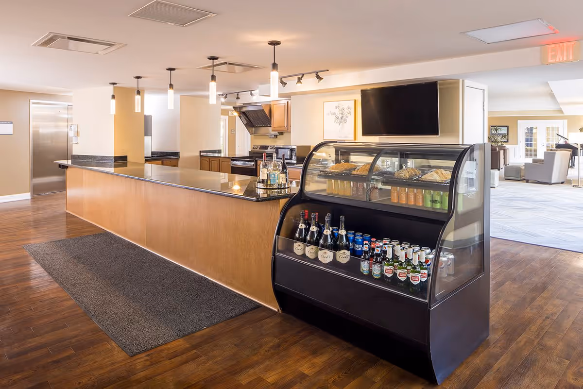 Interior view of a facility's kitchen and common area with a long counter, pendant lights, a display case filled with beverages and pastries, and a seating area visible in the background.