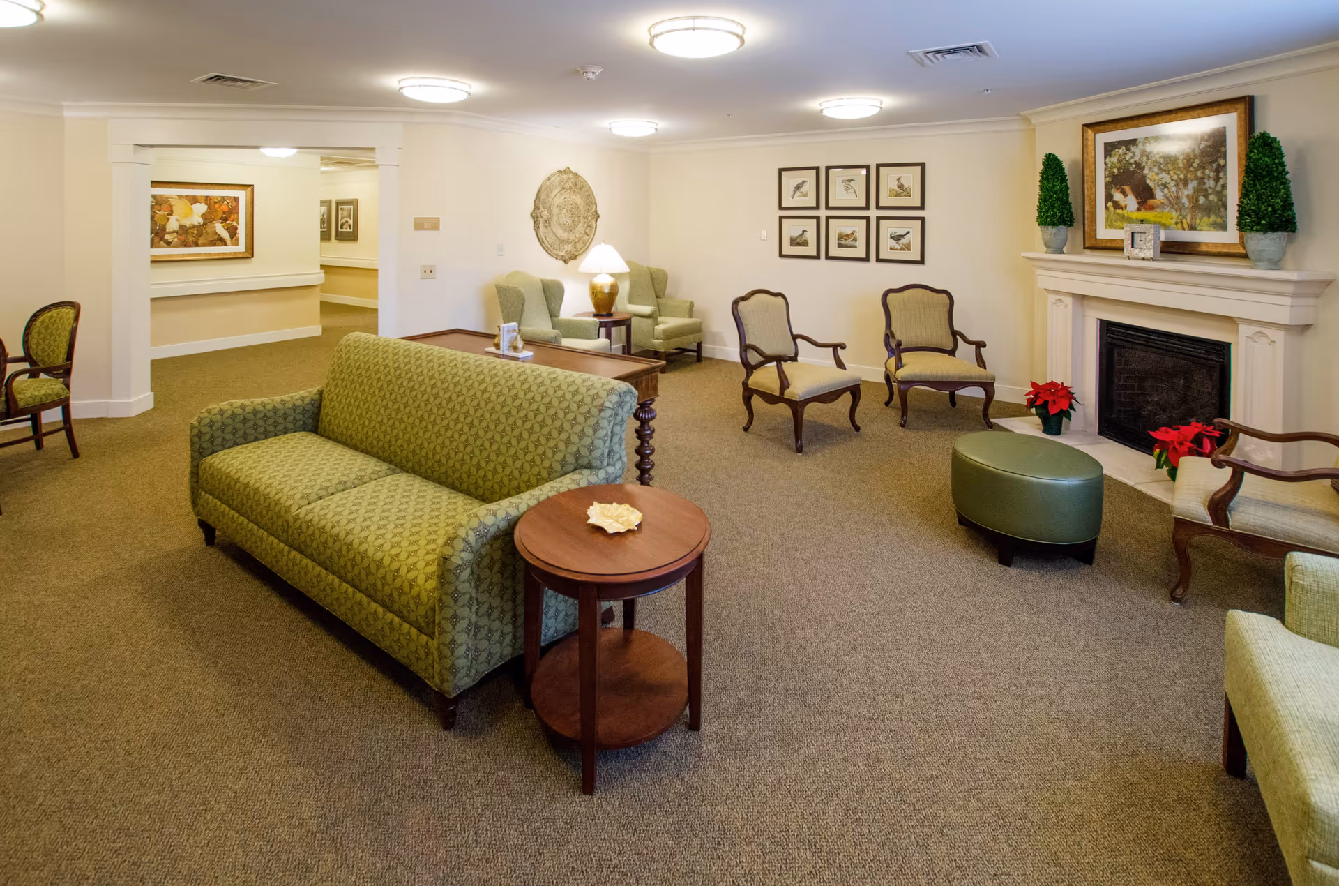 A cozy senior living facility lounge with green upholstered sofas and armchairs arranged around wooden side tables. The room features a fireplace decorated with framed artwork and potted plants. The walls are adorned with framed pictures and a decorative wall hanging. The space is well-lit with ceiling lights and has a carpeted floor.