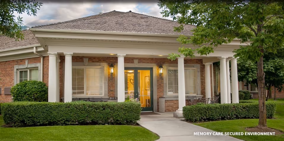 Exterior view of a brick building with white columns and a covered porch entrance, surrounded by neatly trimmed bushes and green grass under a cloudy sky.
