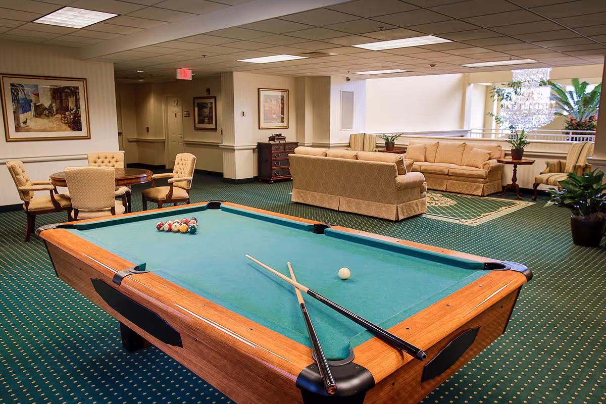 A spacious indoor lounge area featuring a pool table with two cues and a set of billiard balls arranged for a game. In the background, there are beige upholstered sofas and armchairs arranged around a patterned rug, with side tables and potted plants. The room has green carpet with a small dotted pattern, framed artwork on the walls, and a ceiling with recessed lighting.