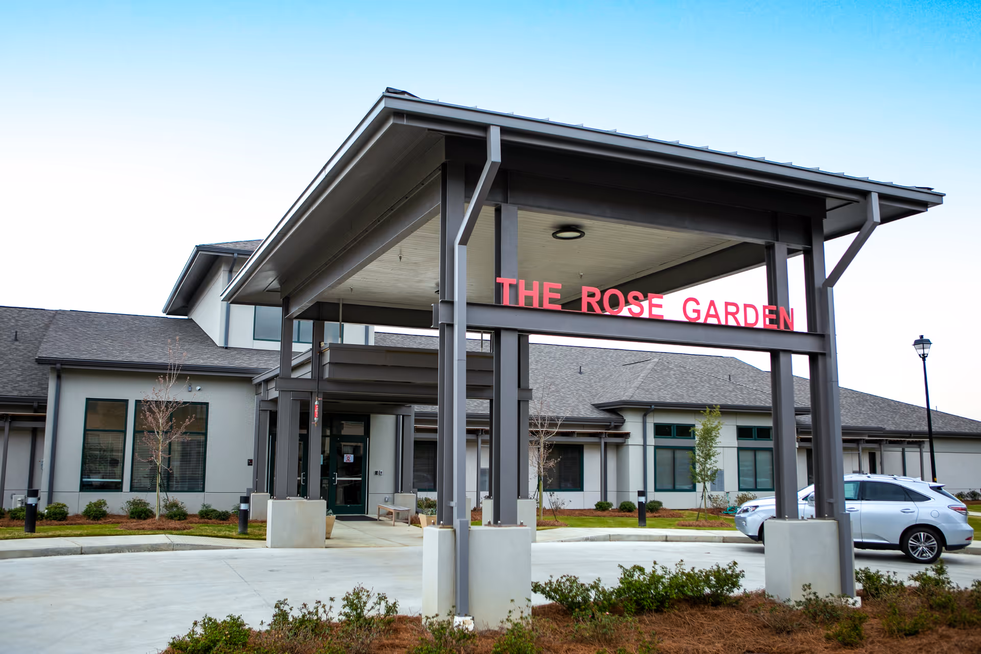 Exterior view of a senior living facility entrance with a covered drop-off area supported by large beams. The sign above the entrance reads 'THE ROSE GARDEN'. There is a silver SUV parked to the right and landscaped bushes and small trees around the building.