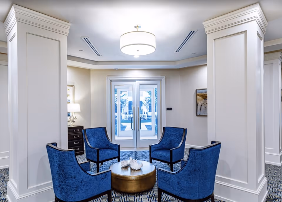 A bright and elegant seating area in a senior living facility with four blue upholstered chairs arranged around a round gold coffee table. The space features white paneled columns, a patterned blue carpet, a modern ceiling light fixture, and double glass doors leading outside. There is a lamp on a dark wooden chest of drawers and a framed artwork on the wall.