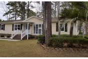 Single-story beige building with a white porch and steps, surrounded by trees and landscaped shrubs.