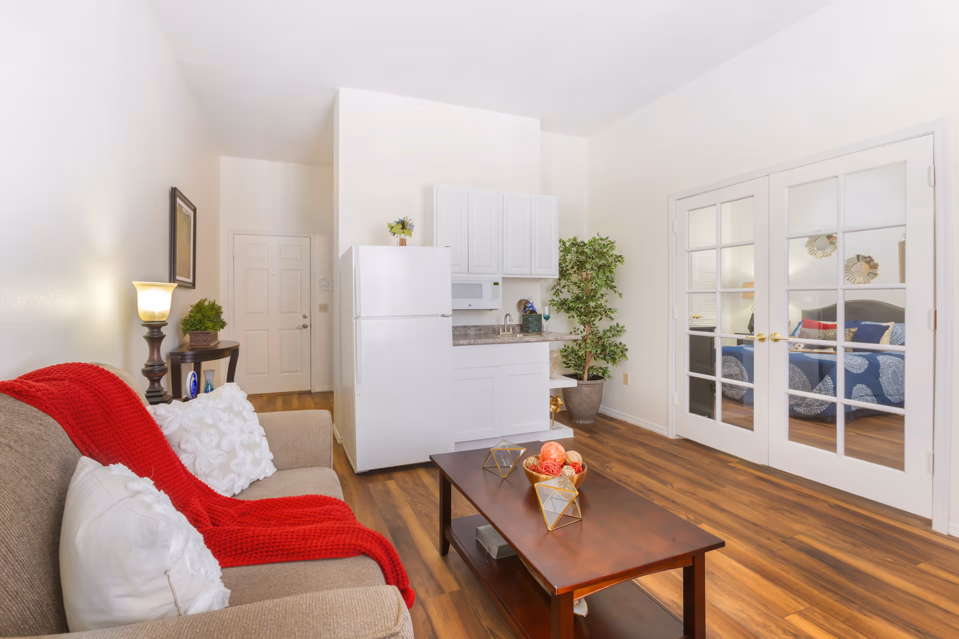 A cozy living room area with a beige sofa draped with a red blanket and white pillows. A wooden coffee table with decorative items sits in front of the sofa. Behind the coffee table is a small kitchenette with white cabinets, a white refrigerator, and a microwave. To the right, glass-paneled double doors reveal a bedroom with a bed covered in blue and white bedding. The room has wooden flooring and a potted plant near the kitchenette.