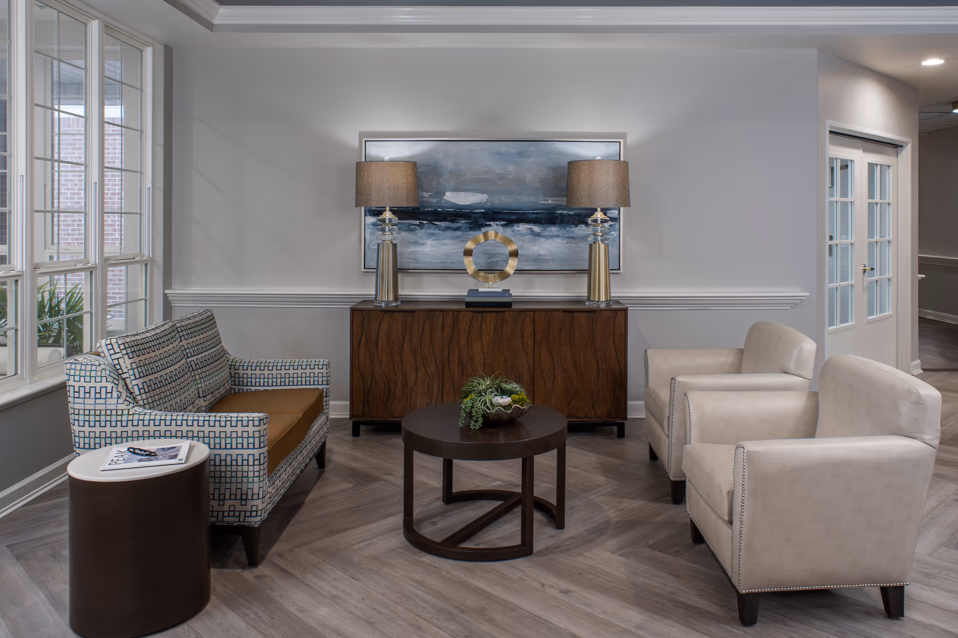 A cozy sitting area in an assisted living facility featuring a patterned loveseat with brown cushions, two beige armchairs, a round dark wood coffee table with a small plant centerpiece, and a wooden sideboard against the wall. On the sideboard are two matching lamps with beige shades and a decorative gold sculpture. Above the sideboard hangs a large abstract painting in shades of blue and gray. The room has light gray walls, large windows on the left, and a door with glass panels on the right.