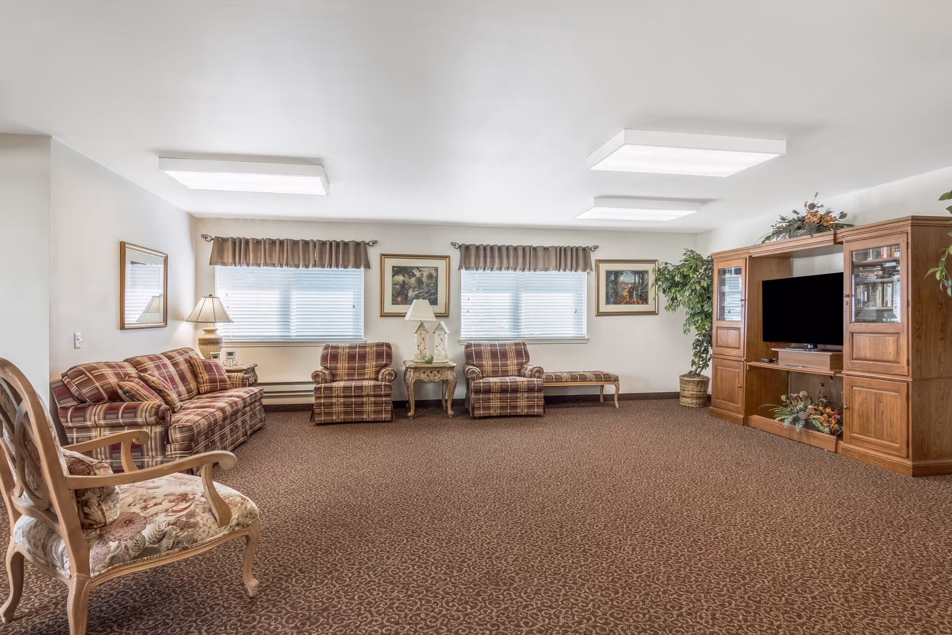 A spacious living room with patterned carpet, a plaid sofa and two matching armchairs, a wooden chair with floral upholstery, a wooden entertainment center with a TV, decorative plants, framed paintings on the walls, and windows with brown valances.