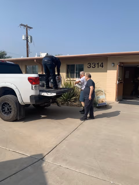 Three men outside a single-story building with the number 3314 on the wall. One man is standing on the open tailgate of a white pickup truck, while the other two men stand on the ground nearby. The building has a beige exterior and an open door leading inside.