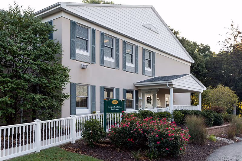 Exterior view of a two-story residential building with beige walls and white trim, featuring multiple windows with gray shutters. A white fence runs along the front, with landscaped bushes and flowering plants. A green sign near the entrance reads '1803 Independent Living Residences'. Trees and greenery surround the building.