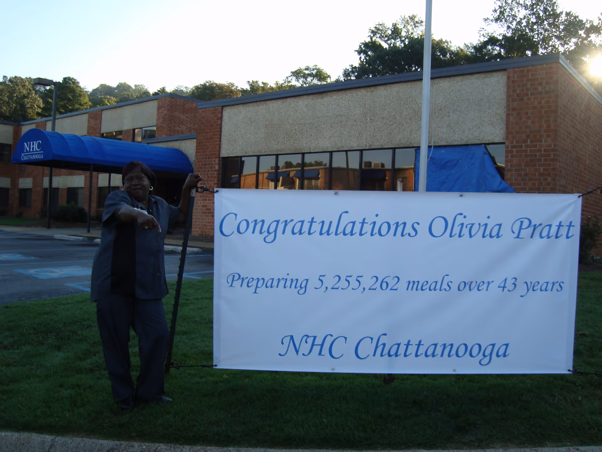 A woman stands outside a brick building with a blue awning labeled 'NHC Chattanooga.' She is pointing towards a large white banner that reads, 'Congratulations Olivia Pratt, Preparing 5,255,262 meals over 43 years, NHC Chattanooga.'