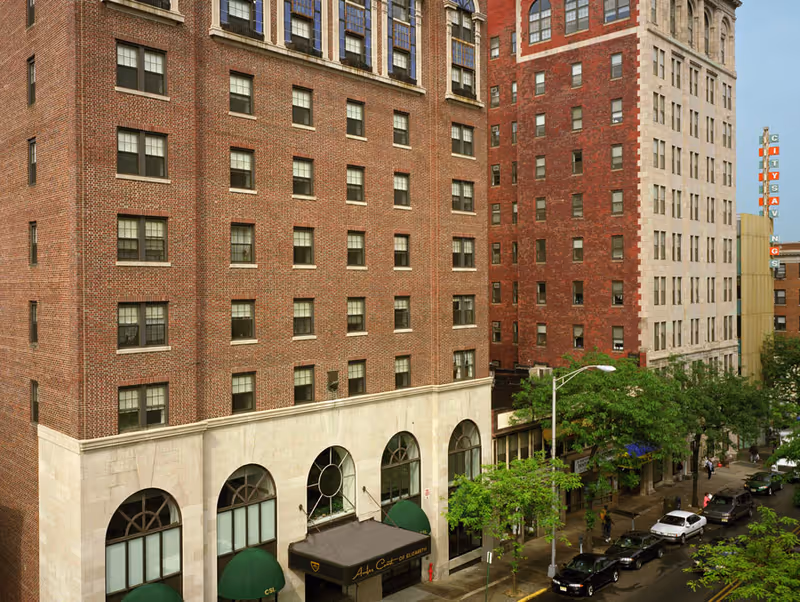 Exterior view of a multi-story brick building with large arched windows on the ground floor and green awnings. The building is located on a city street with trees, parked cars, and pedestrians. A sign on the awning reads 'Amber Court of Elizabeth'.