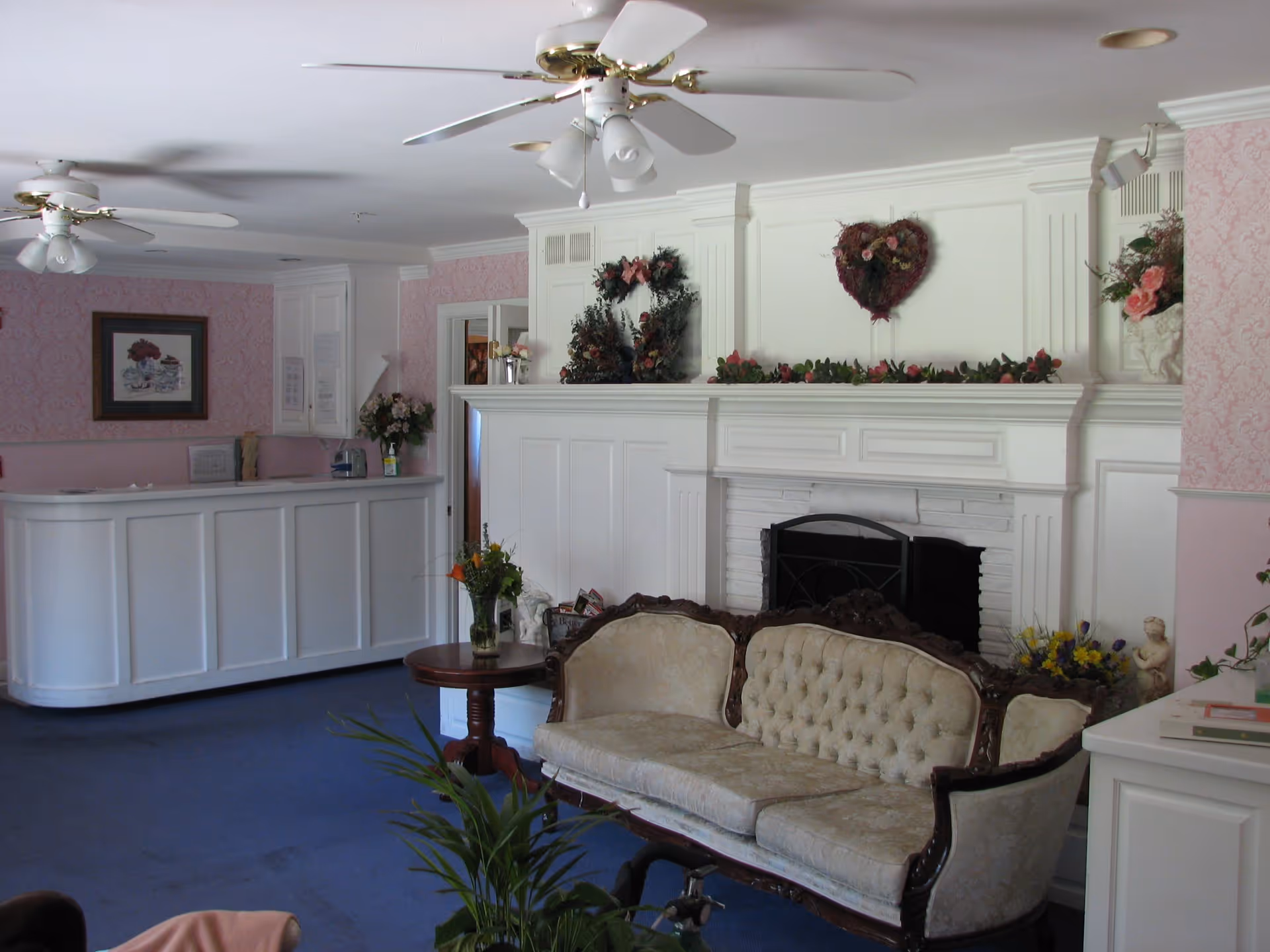 A cozy assisted living facility common area with a vintage upholstered sofa, a small wooden side table with a vase of flowers, a white fireplace decorated with floral wreaths and garlands, pink wallpaper, ceiling fans, and a white reception desk in the background.