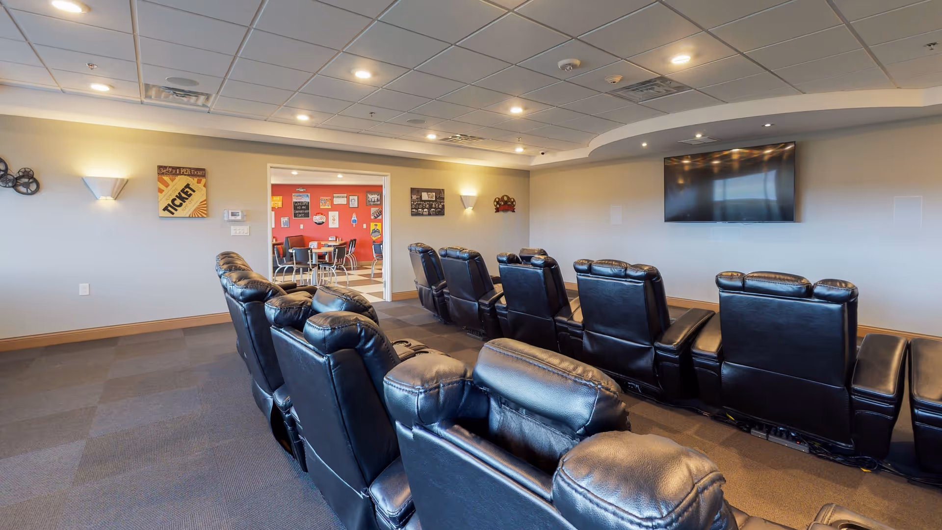 A small theater room with two rows of black leather recliner chairs facing a large flat-screen TV mounted on the wall. The room has a carpeted floor, recessed lighting in the ceiling, and light-colored walls. Through an open doorway, a red-walled room with tables and chairs is visible.