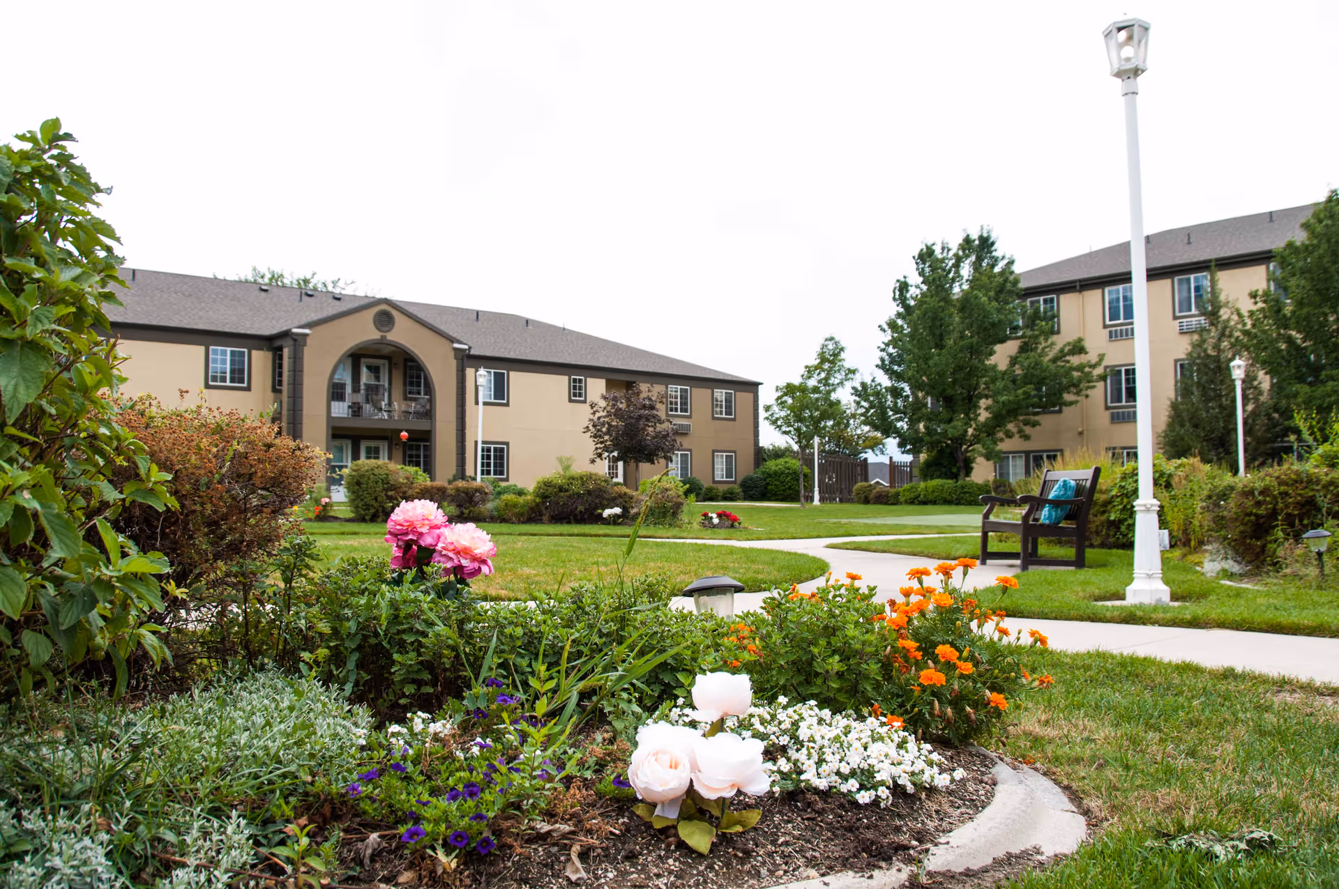 A landscaped garden area with colorful flowers and green shrubs in the foreground, a curved concrete pathway, benches, and two beige multi-story residential buildings in the background under an overcast sky.