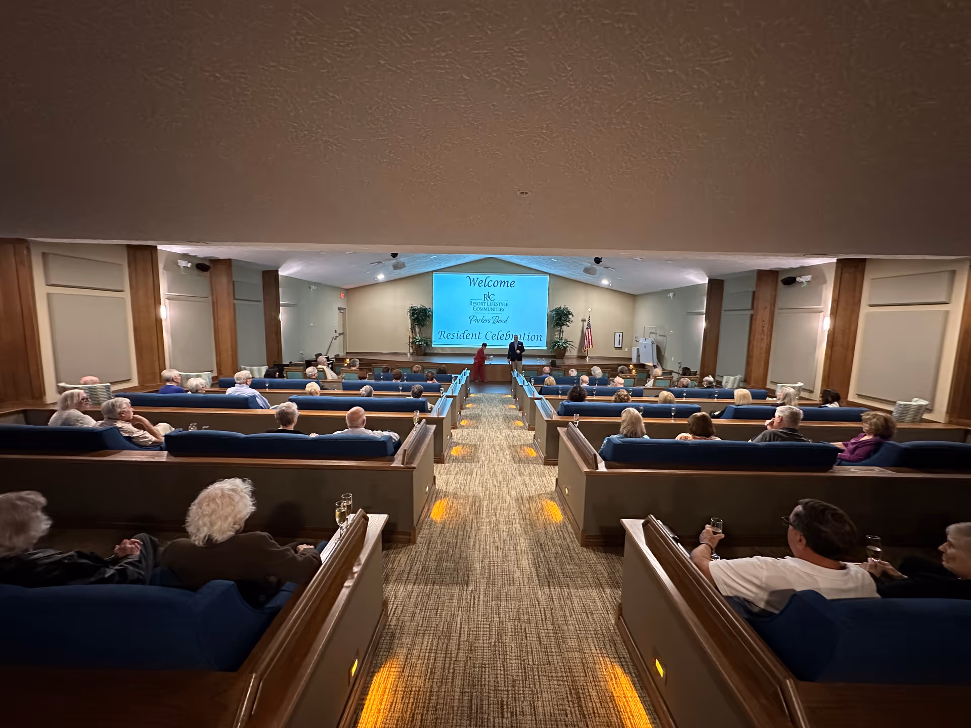 Interior view of a community room with rows of blue cushioned benches filled with elderly residents facing a stage with a large screen displaying a welcome message for Parkers Bend Resident Celebration. The room has beige walls, wood paneling, and carpeted floor with aisle lighting.