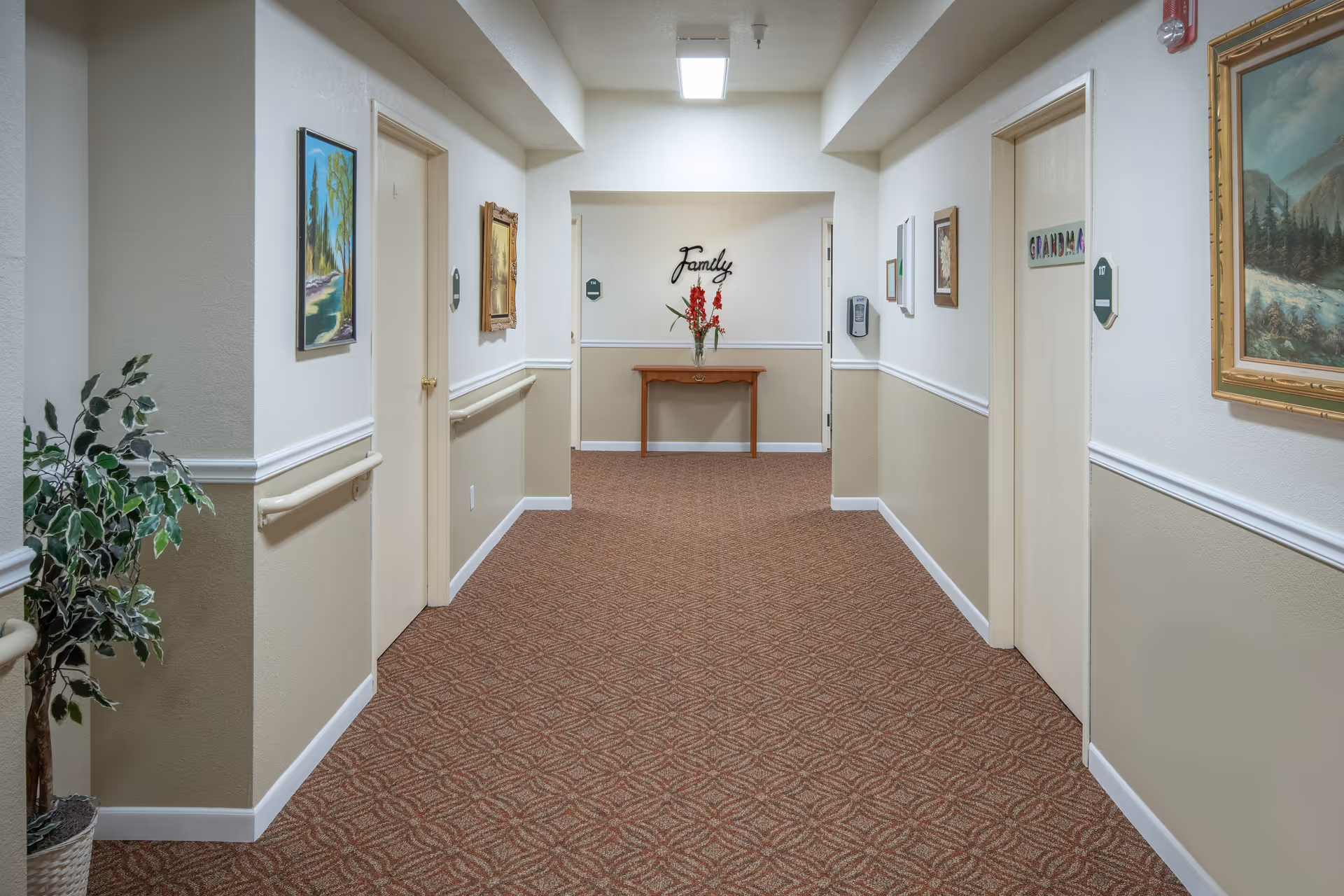 A carpeted hallway in an assisted living facility with beige and white walls, several closed doors, framed paintings on the walls, a potted plant on the left, and a small wooden table with a vase of red flowers at the end under a wall decoration that says 'Family'.