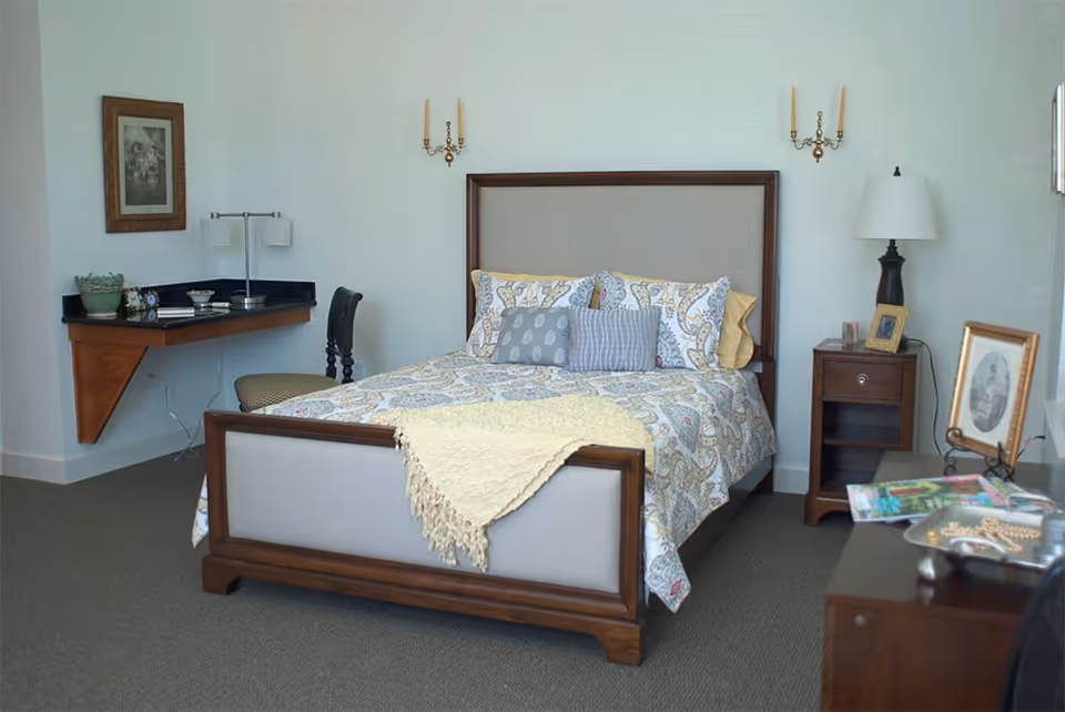 A neatly made bed with a wooden frame and patterned bedding in a bedroom. There are two wall-mounted candle holders above the bed, a nightstand with a lamp and picture frame on the right, and a small desk with a chair and lamp on the left. A dresser with magazines and decorative items is visible in the foreground.