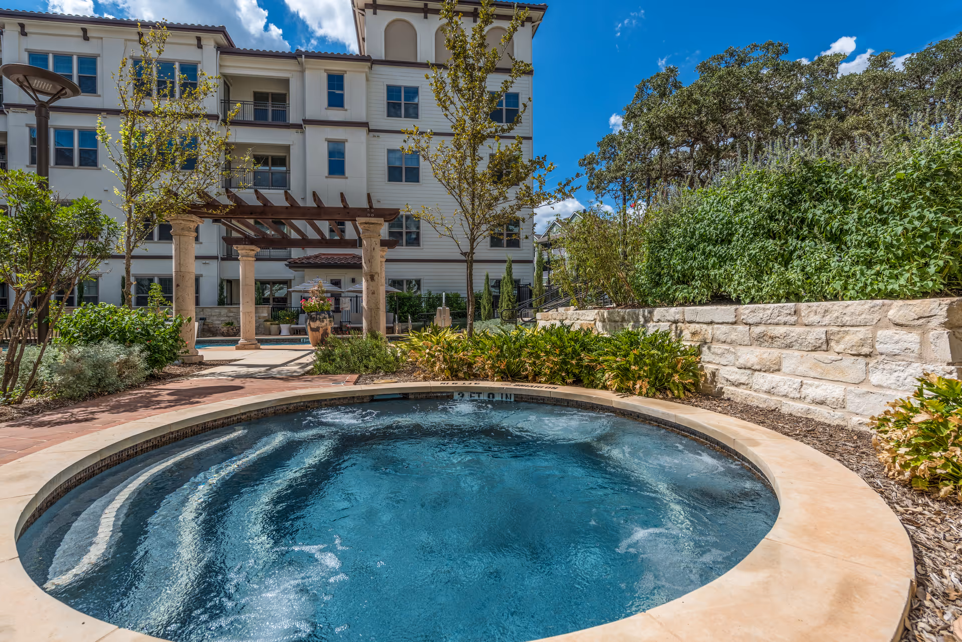 Circular bubbling outdoor spa in a landscaped courtyard with a pergola and a multi-story building in the background.