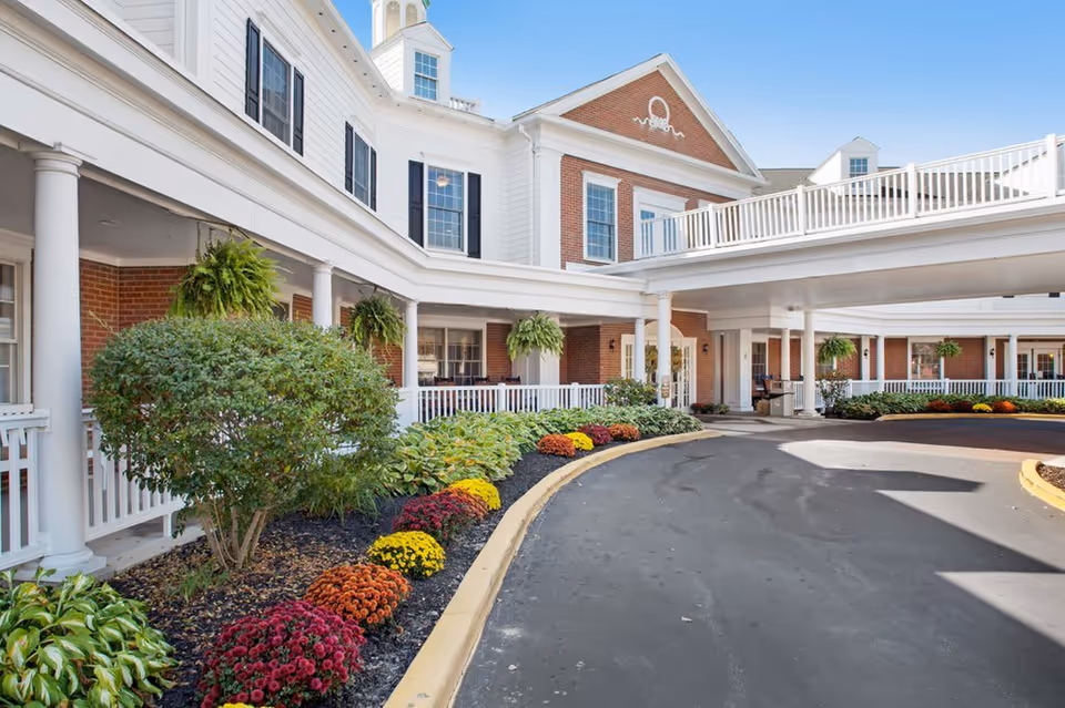Exterior view of Poland Village Assisted Living facility showing a curved driveway, landscaped flower beds with colorful flowers and shrubs, white railings, columns, and a brick and white-paneled building under a clear blue sky.