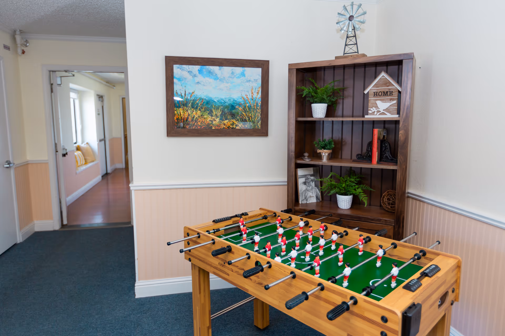 A foosball table in a room with a wooden bookshelf holding plants, a small windmill decoration, and a wooden house-shaped sign that says 'HOME'. A colorful landscape painting hangs on the wall above the foosball table. The room has light-colored walls with a chair rail and wainscoting, and a hallway with windows and seating is visible in the background.