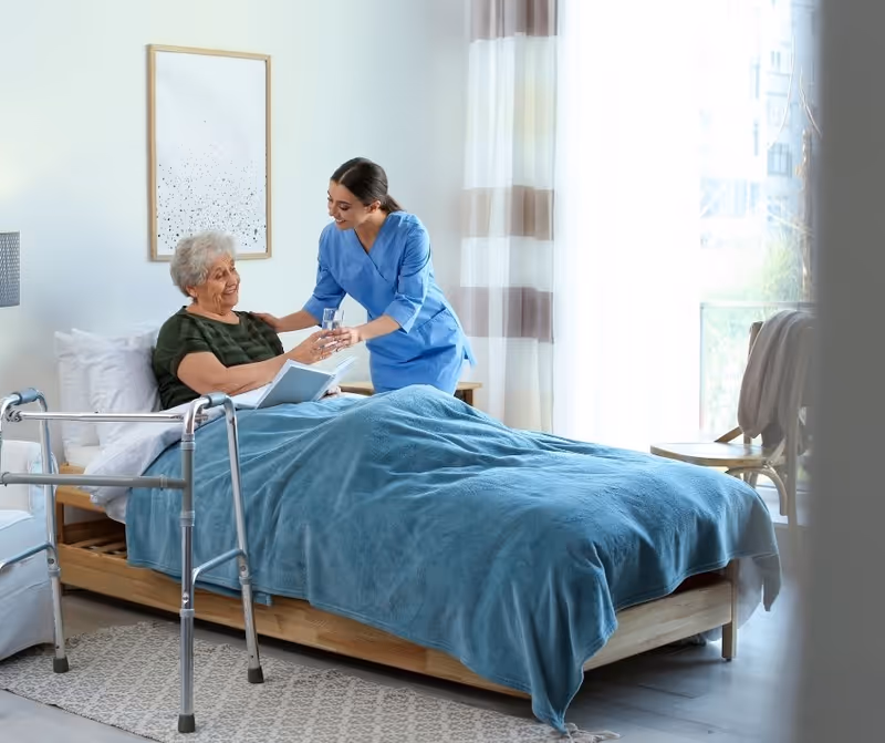 A caregiver in blue scrubs handing a glass of water to an elderly woman sitting up in bed covered with a blue blanket in a bright, cozy bedroom with a walker nearby and sheer curtains letting in natural light.