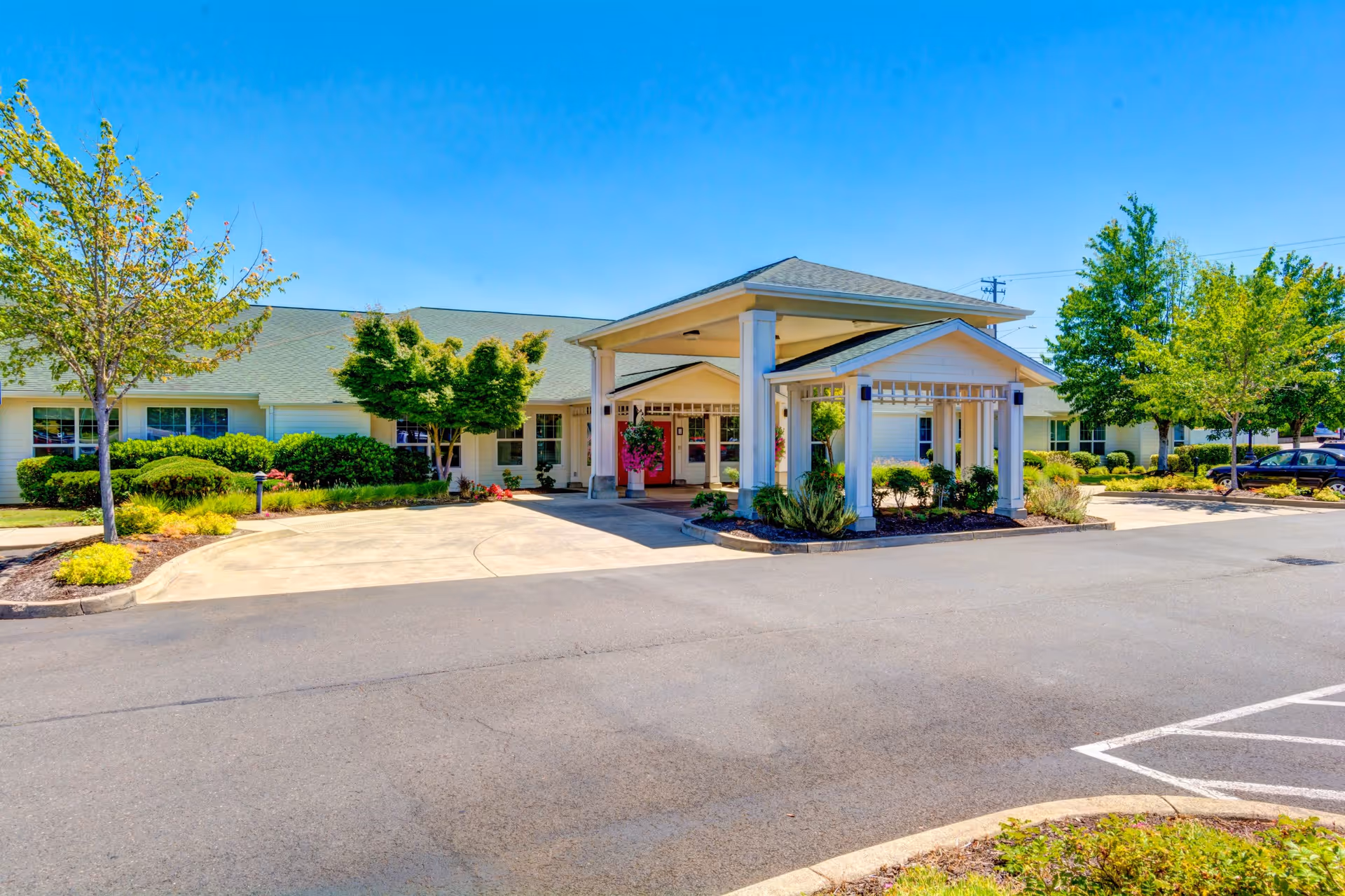 Exterior view of Callahan Court Memory Care facility showing a single-story building with a covered entrance, surrounded by landscaped greenery and trees under a clear blue sky.