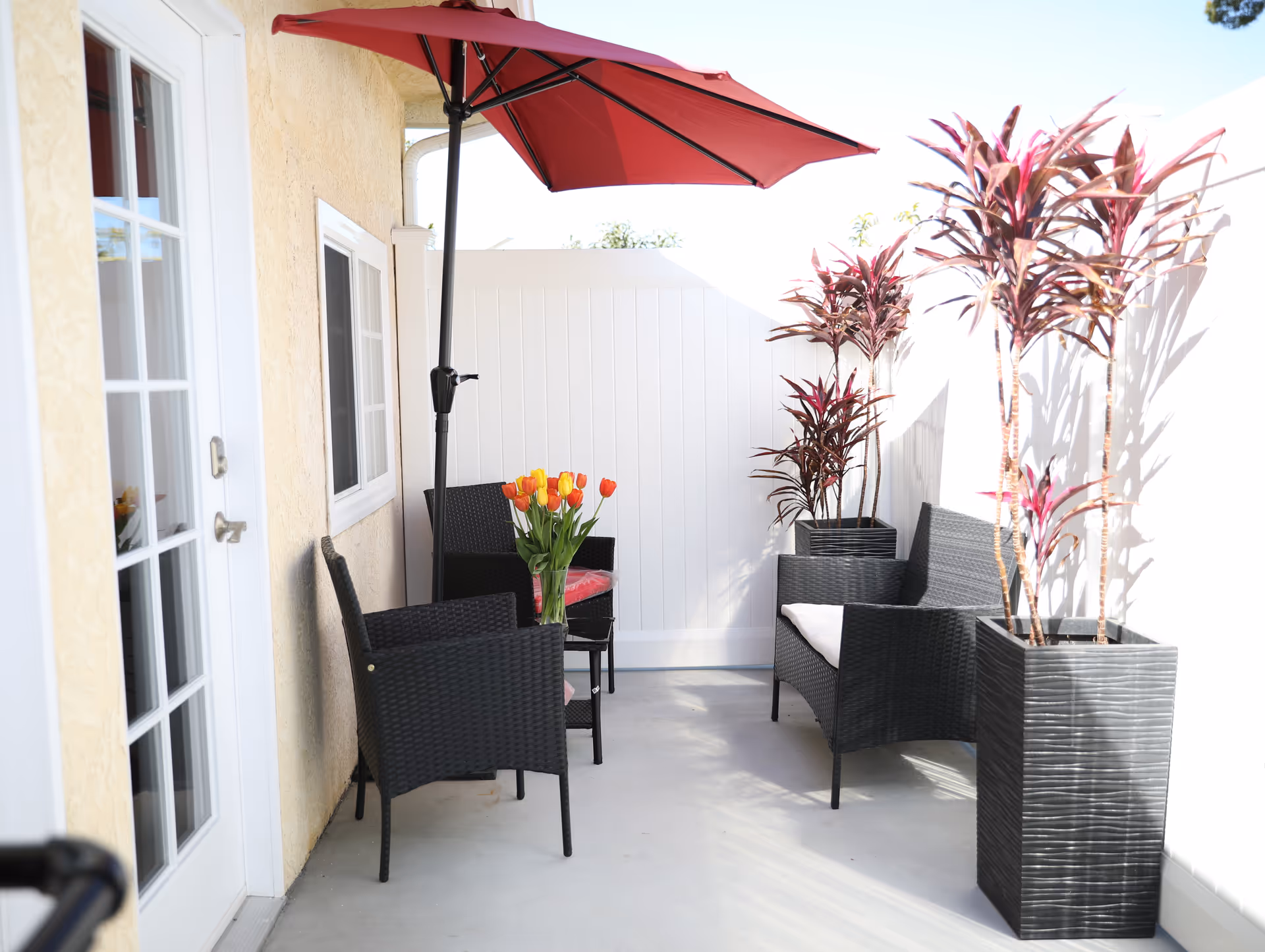 Small outdoor patio with black wicker chairs, a red umbrella, potted plants, and a vase of tulips on a table.