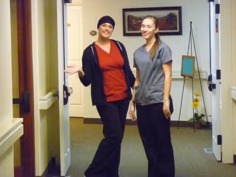 Two women standing in a hallway of a senior living facility. One woman is wearing a red top with a black jacket and head covering, gesturing with her hand, while the other woman is wearing a gray scrub top and black pants. The hallway has beige walls, a framed picture, a small easel, and a potted plant with yellow flowers.