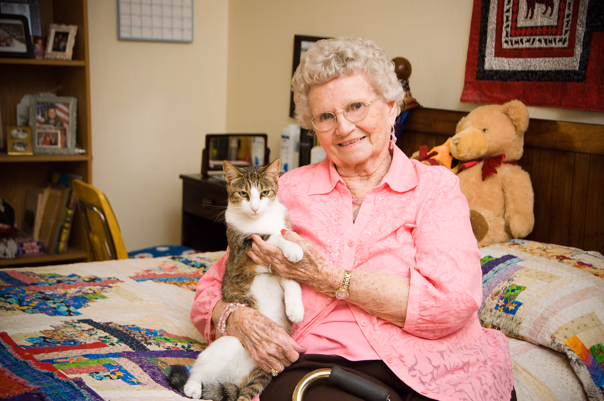 An elderly woman with curly white hair and glasses sits on a bed holding a tabby and white cat. She is smiling and wearing a pink blouse. Behind her on the bed is a large teddy bear and a colorful quilt. A wooden bookshelf and a desk with personal items are visible in the background.