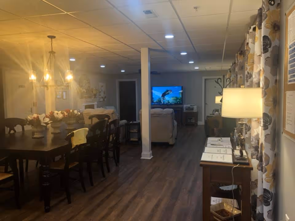 Interior common area with a dining table and chandelier in the foreground and a seating area with a TV and lamps in the background.