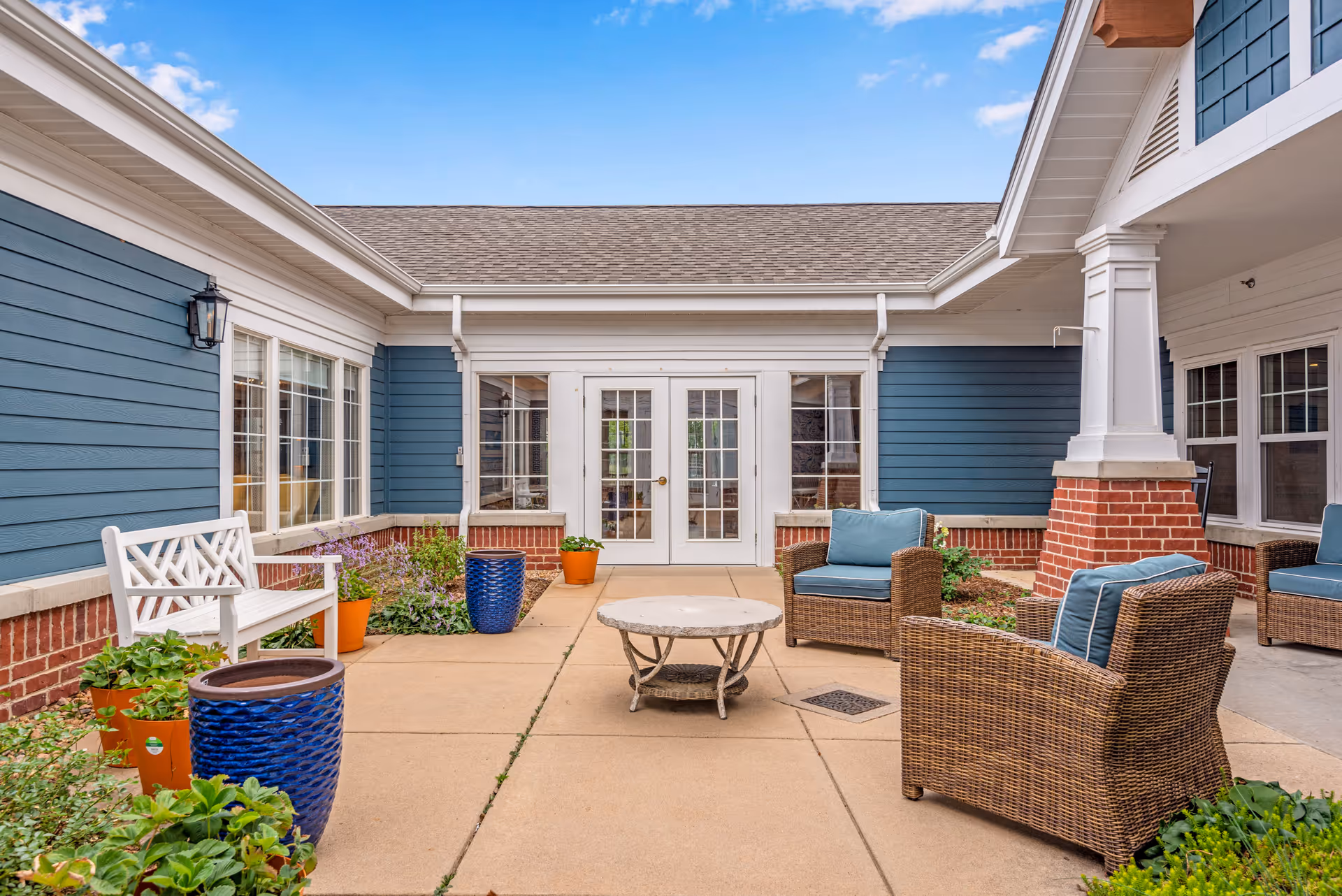 Outdoor patio area at Balfour Cherrywood featuring wicker chairs with blue cushions, a white bench, a round coffee table, potted plants, and blue siding with white trim under a clear blue sky.