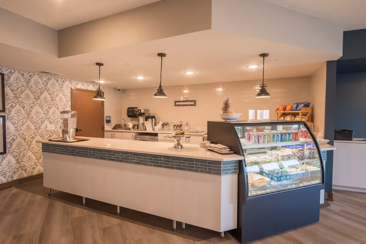 Interior view of a cafe counter featuring a display case with beverages and snacks, a coffee station, and decorative wallpaper.
