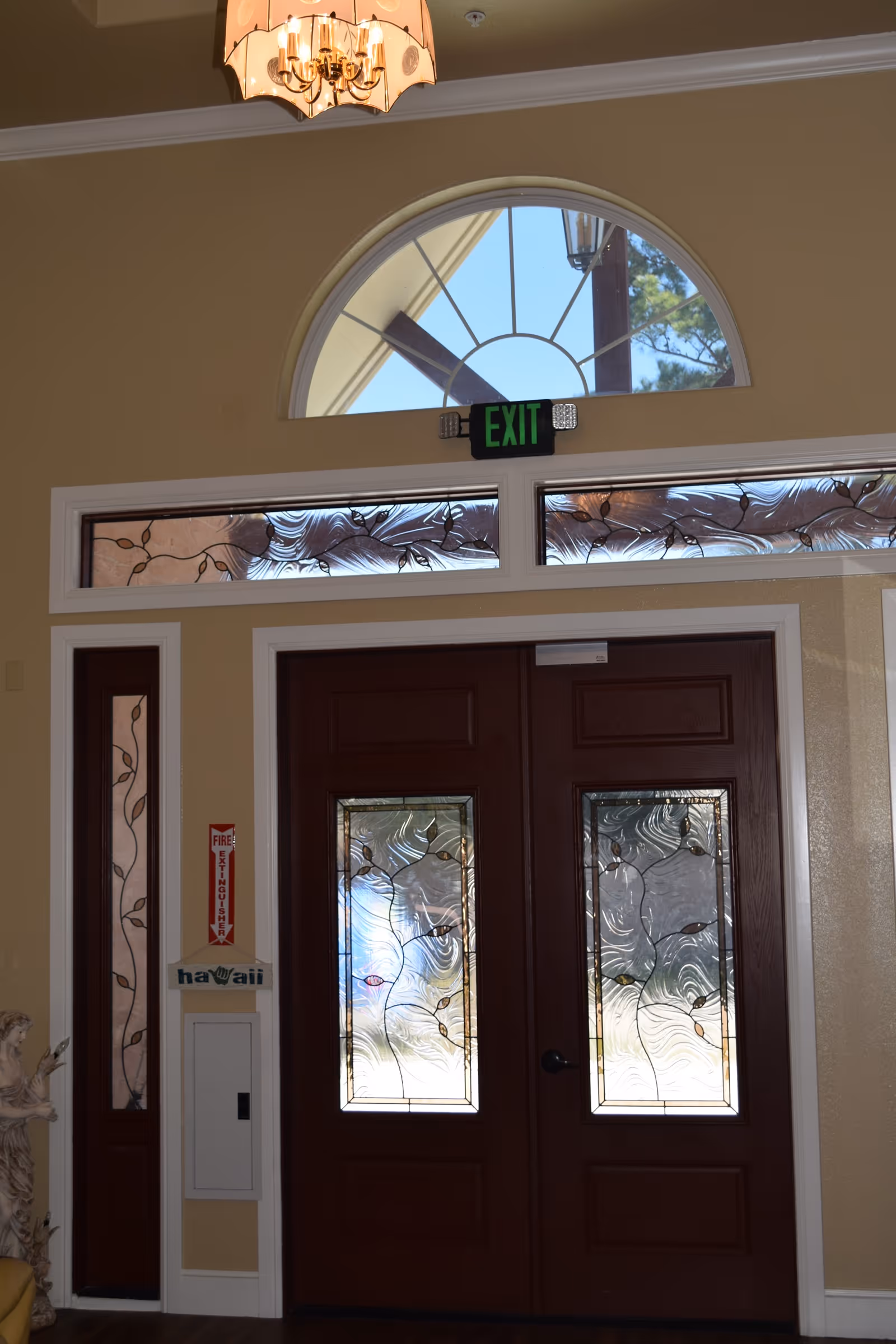 Interior view of a facility entrance featuring double wooden doors with decorative frosted glass panels. Above the doors is a semi-circular window and a green exit sign. The walls are painted beige, and a chandelier hangs from the ceiling. A fire extinguisher sign and a small statue are visible to the left of the doors.