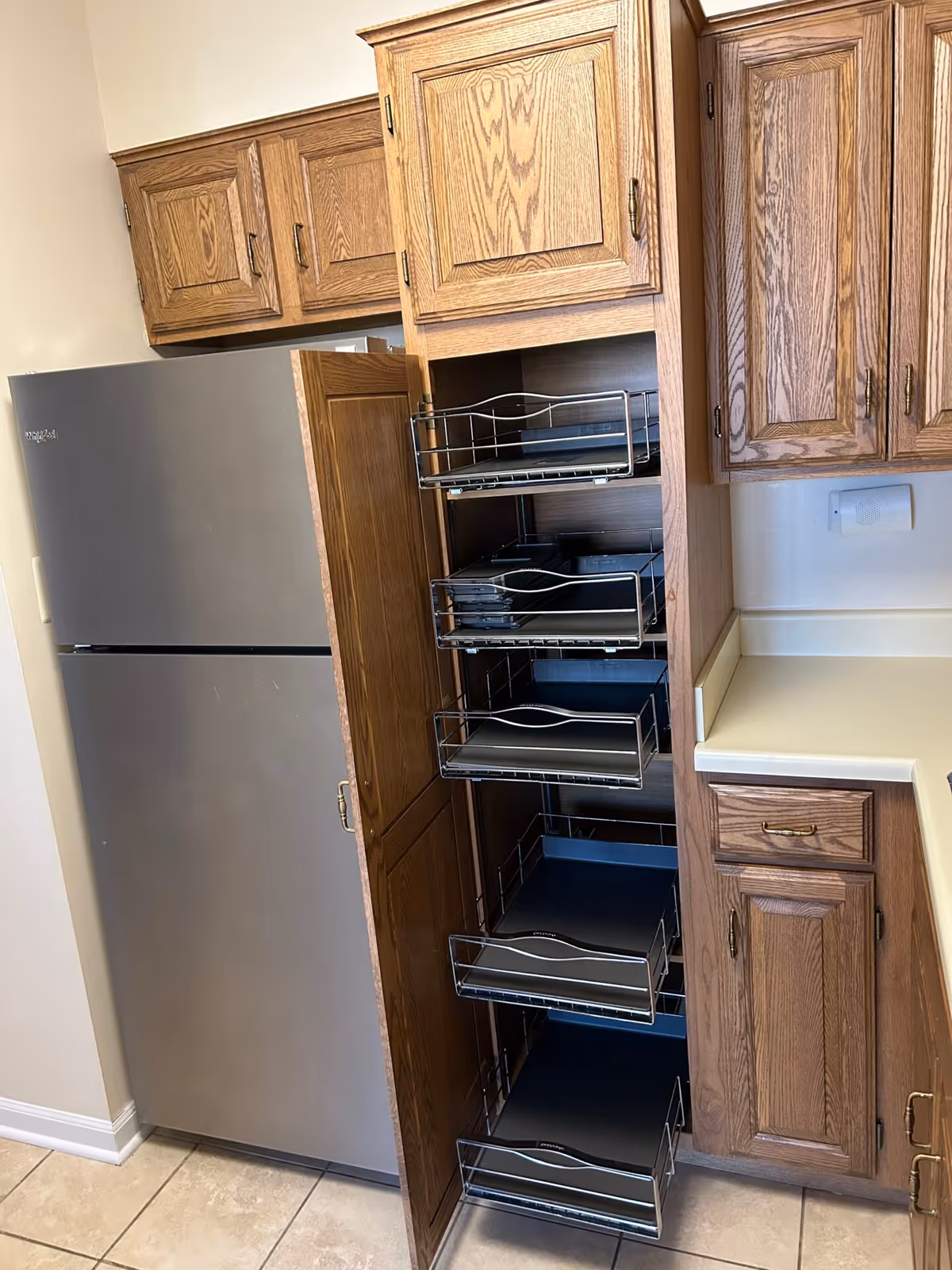 Tall wooden pull-out pantry with multiple metal sliding baskets beside a stainless steel refrigerator and oak kitchen cabinets.