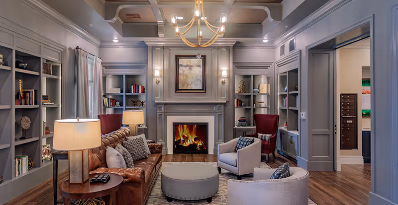 Elegant communal living room with a leather sofa, armchairs, round ottoman, fireplace, built-in bookshelves and chandelier.