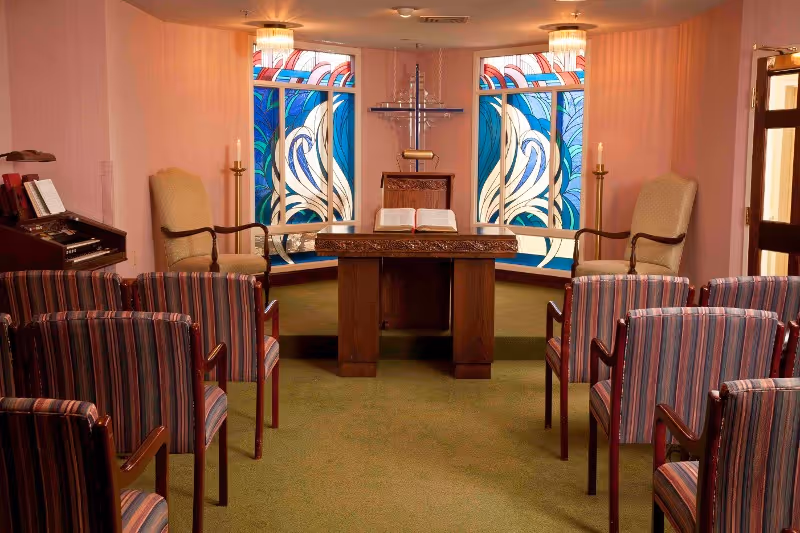 Interior of a small chapel or prayer room with rows of striped chairs facing a wooden altar with an open book. Behind the altar are two stained glass windows with blue, white, and red patterns, and a cross mounted on the wall. Two tall candles are on either side of the altar, and two upholstered armchairs are positioned near the windows. The room has soft pink walls and green carpet.