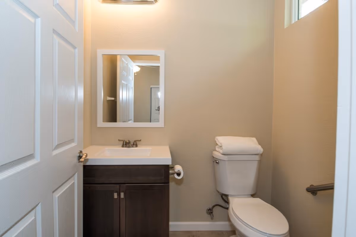 Small bathroom with a white toilet topped with folded white towels, a dark wood vanity with a white sink, a silver faucet, and a rectangular mirror above it. The walls are beige, and there is a white door partially open on the left side.