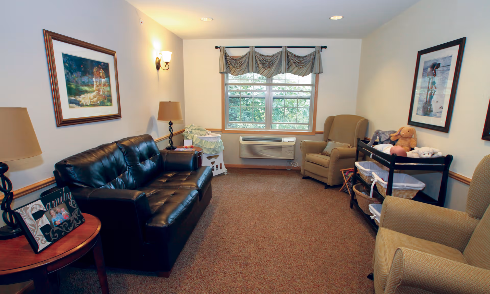 Cozy sitting room with a black leather sofa, upholstered armchairs, a window with an air conditioner, lamps, and framed artwork.