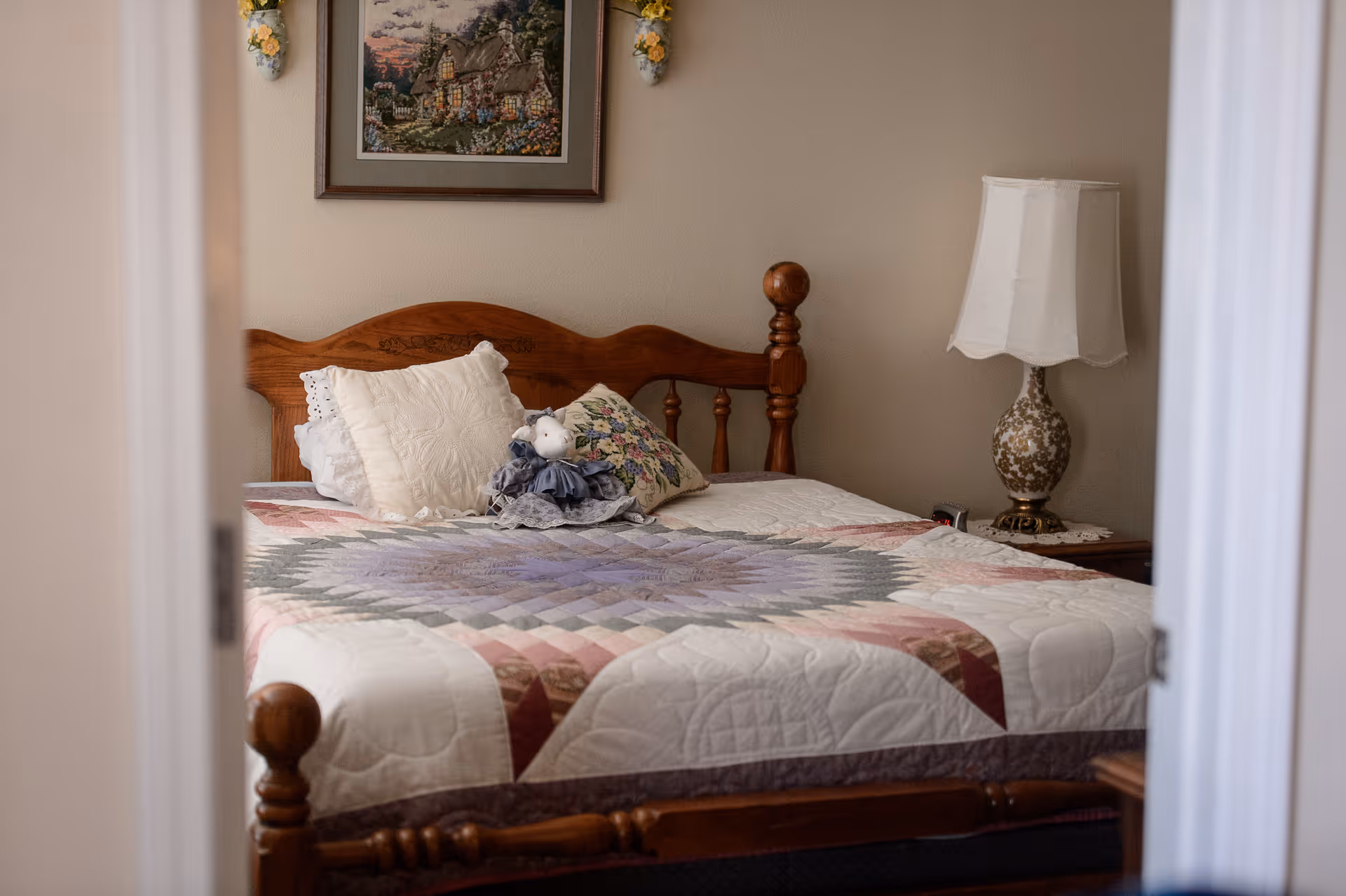 A cozy bedroom with a wooden bed dressed in a patchwork quilt, decorative pillows, a small doll, and a bedside lamp.
