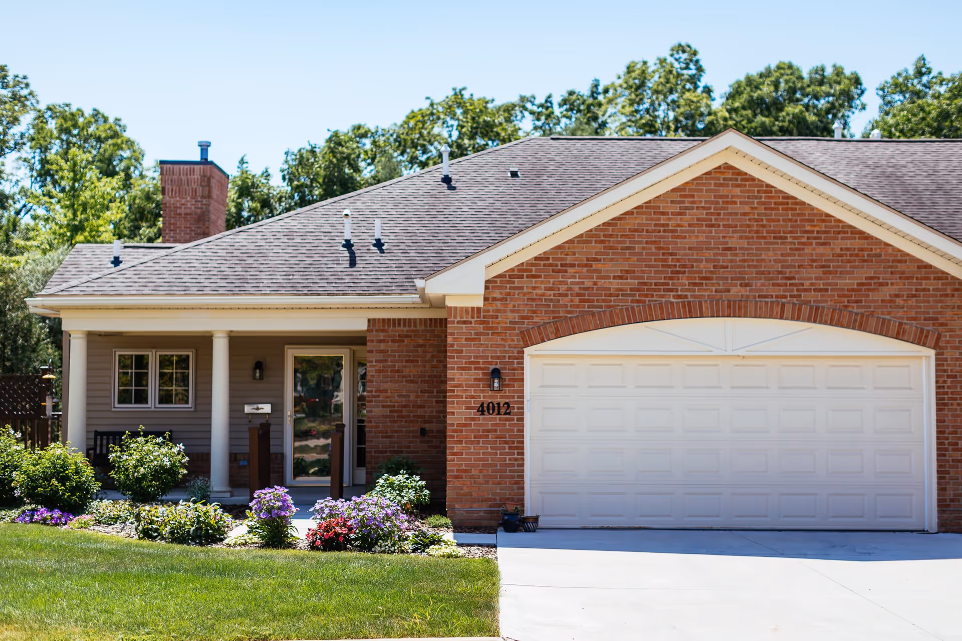 Front exterior view of a single-story brick and siding house with a white garage door, a small porch with two white columns, a glass front door, and a well-maintained garden with green shrubs and colorful flowers in front. Trees are visible in the background under a clear blue sky.