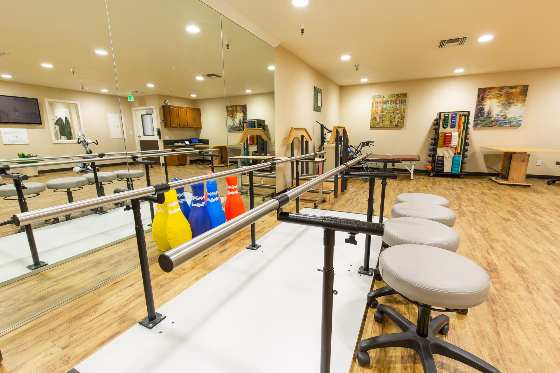 A bright therapy or exercise room with wooden floors and beige walls. The room features parallel bars for walking exercises, several round stools with wheels, colorful bowling pins, exercise equipment, and two large paintings on the walls. A large mirror covers one wall, reflecting the room's contents.