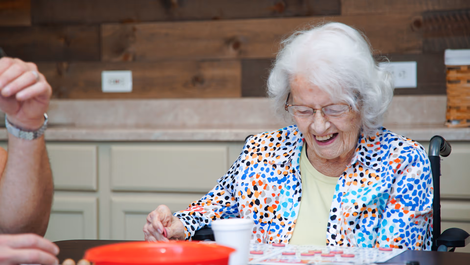 An elderly woman with white hair and glasses, wearing a colorful spotted shirt, is sitting at a table playing a board game and smiling. A person's arm is partially visible on the left side. The background shows a kitchen counter with cabinets and a wooden wall.