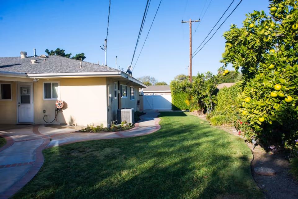 Outdoor view of a single-story building with a beige exterior, a gray shingled roof, and a white door. There is a green lawn with a curved concrete pathway bordered by red bricks. On the right side, there are green bushes and trees with yellow fruit. Power lines and a utility pole are visible against a clear blue sky.
