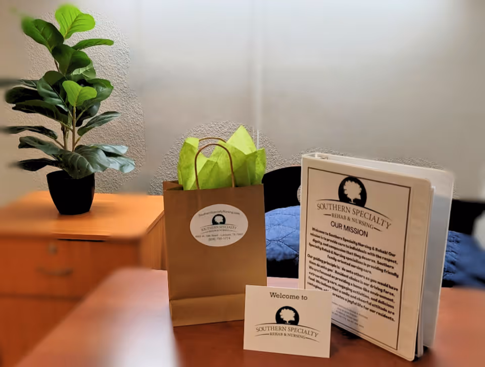 A table with a potted plant, a paper gift bag with green tissue paper, and a binder and welcome card labeled 'Southern Specialty Rehab & Nursing'.