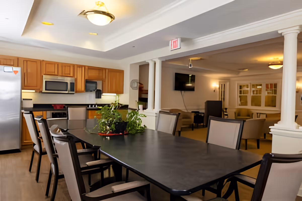 A spacious kitchen and dining area in a senior living facility with a long black dining table surrounded by cushioned chairs. The kitchen features wooden cabinets, a stainless steel refrigerator, microwave, and dishwasher. In the background, there is a living room area with armchairs, a wall-mounted TV, and soft lighting.