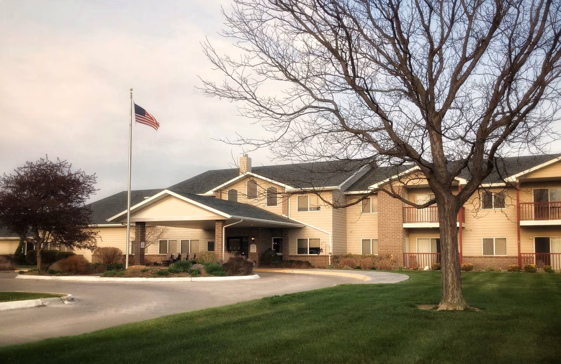 Exterior view of a two-story senior living facility building with beige siding and brick accents. There is a covered entrance with a driveway in front, an American flag on a flagpole, and a large leafless tree on a well-maintained green lawn.