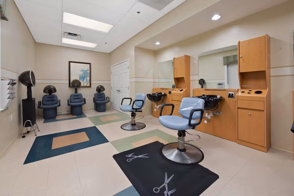 Interior view of a hair salon area in a senior living facility with two salon chairs in front of mirrors and sinks on the right side, and three hair drying chairs with hooded dryers along the back wall. The floor has a patterned design with colored tiles, and there is a framed picture on the back wall.