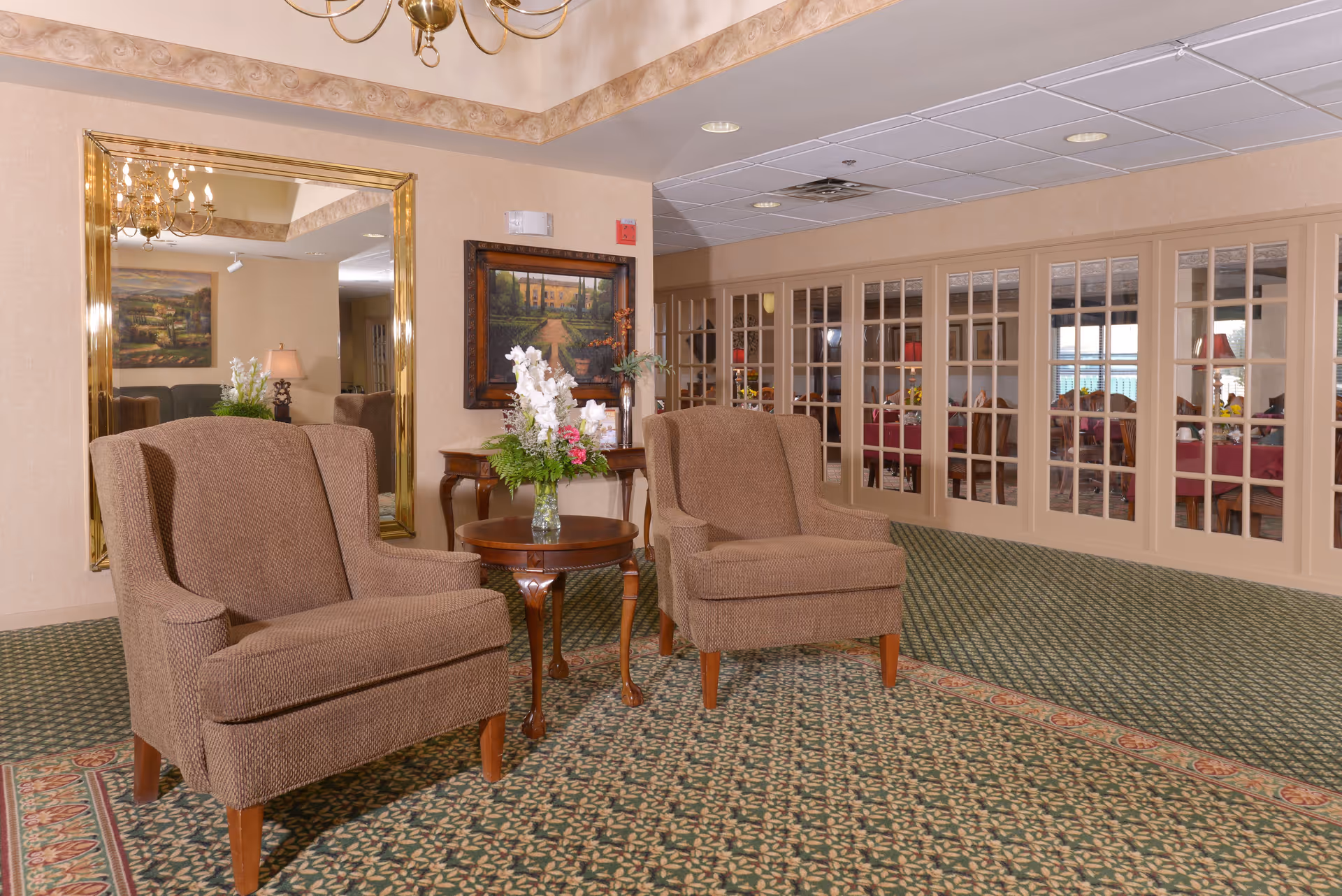 A cozy seating area in a senior living facility with two brown upholstered armchairs positioned around a small wooden table holding a vase of white and pink flowers. Behind the chairs is a large gold-framed mirror reflecting part of the room, and a framed painting on the wall. To the right, there are glass-paneled doors leading to a dining area with tables covered in red tablecloths and chairs.