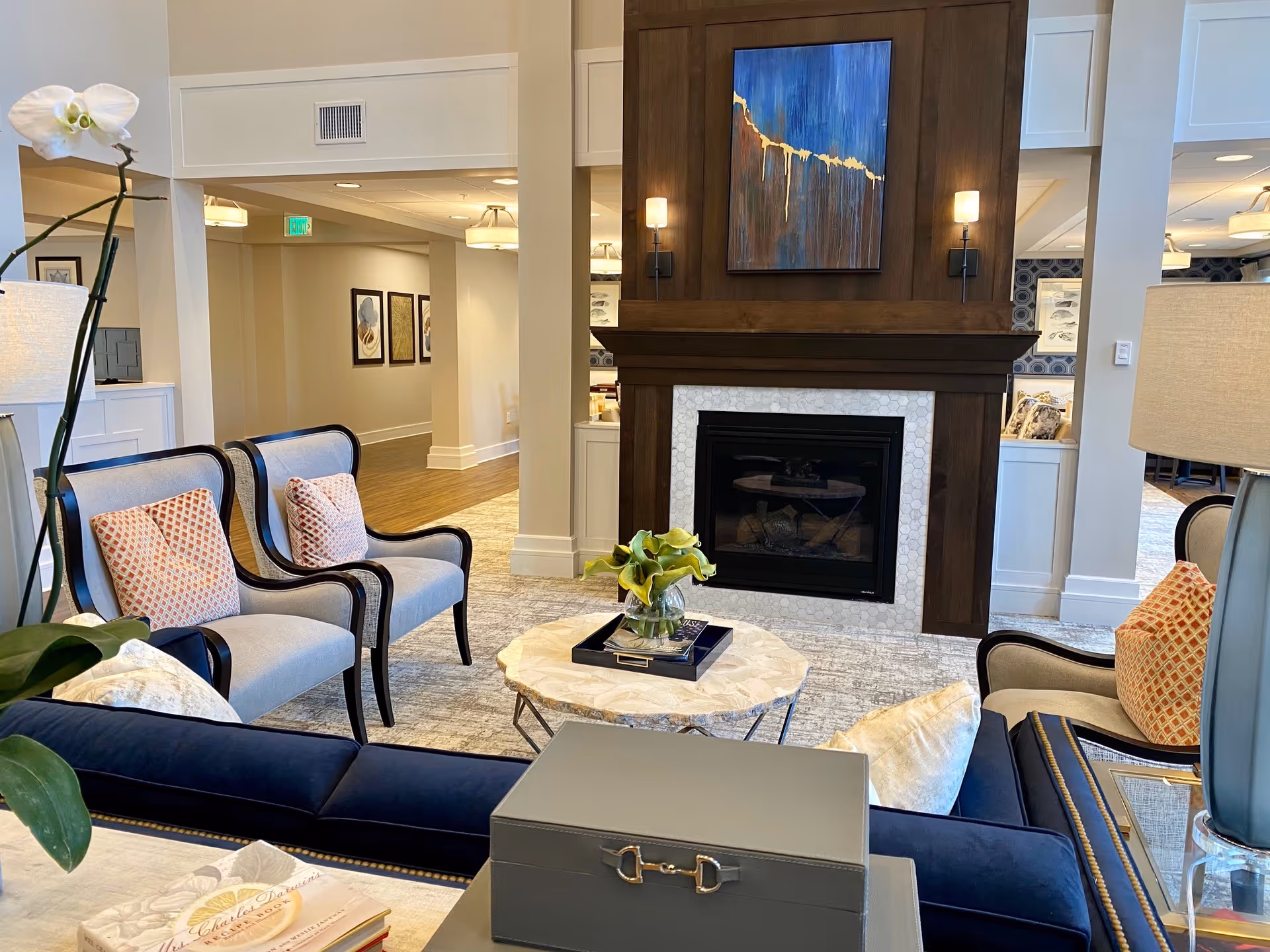 A cozy communal lounge with upholstered chairs, a marble-topped coffee table, and a fireplace topped by artwork.